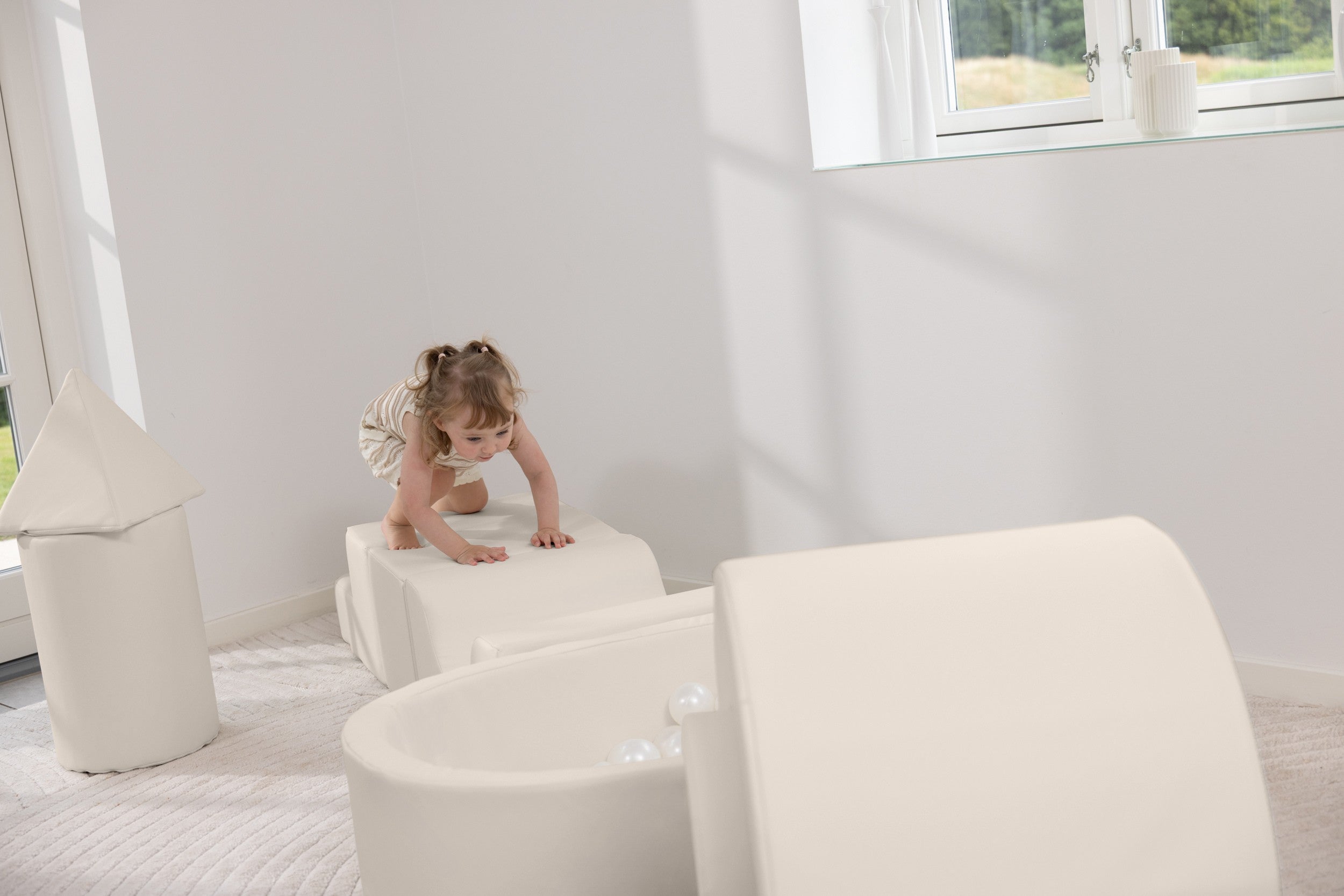 Child playing on a white ottoman in a room with white furniture and decor.